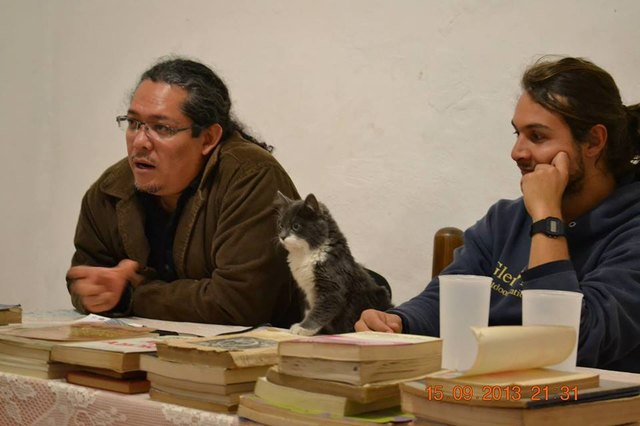 Kitten between to people at table covered with books.