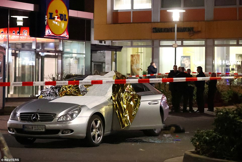 A Mercedes Benz was left damaged with glass covering the floor as police look on at the crime scene in Hanau last night